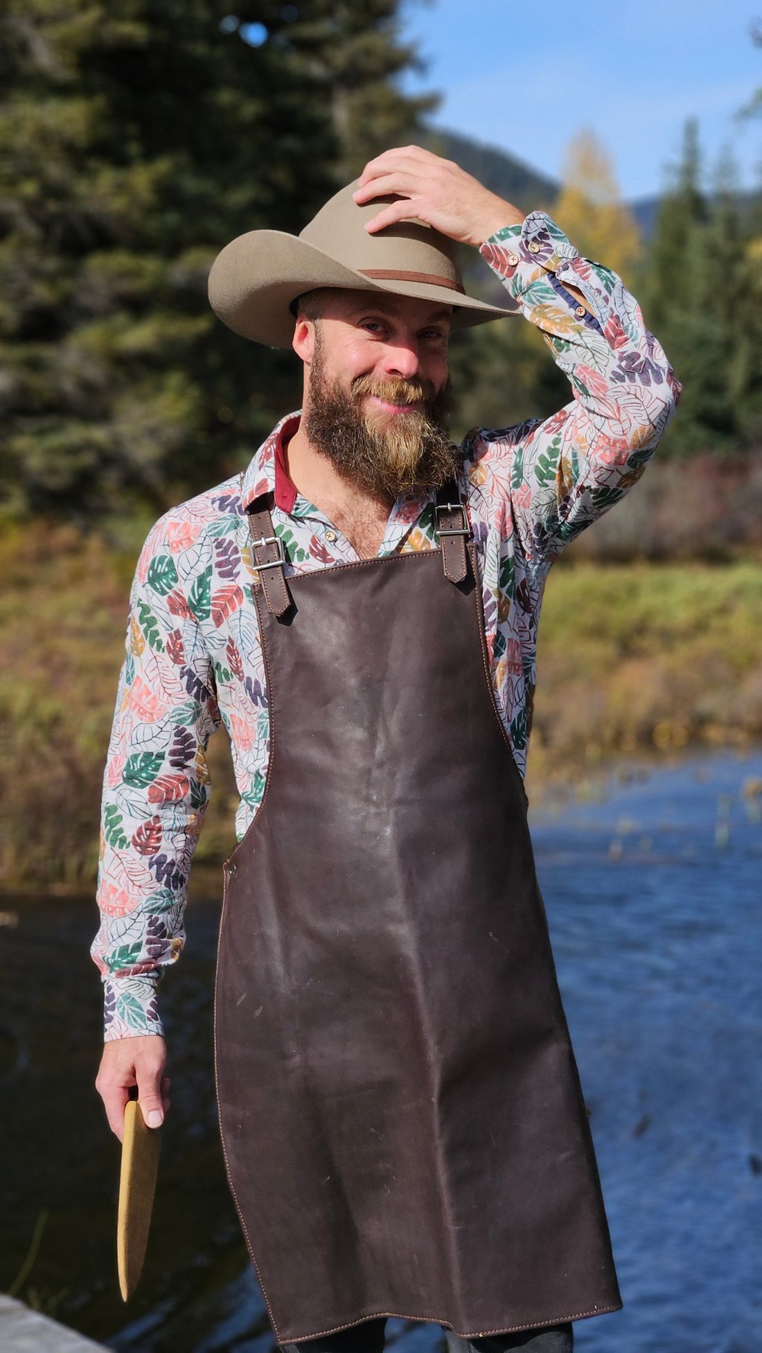 Chef Hugh Flint of Whistler Living Kitchen in a leather apron and felt hat beside a river