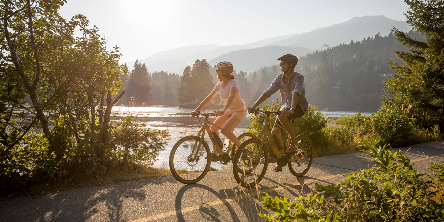 Two riders on e-bikes on a lakeside path in Whistler in evening light