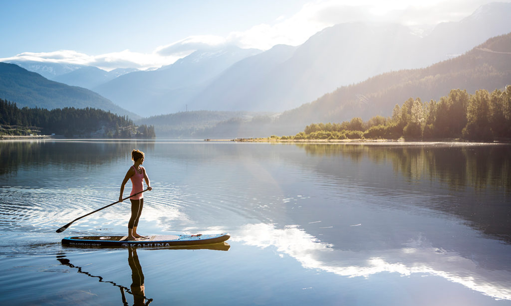 A paddleboarder on the calm water of Alta Lake with Whistler and Blackcomb mountains behind