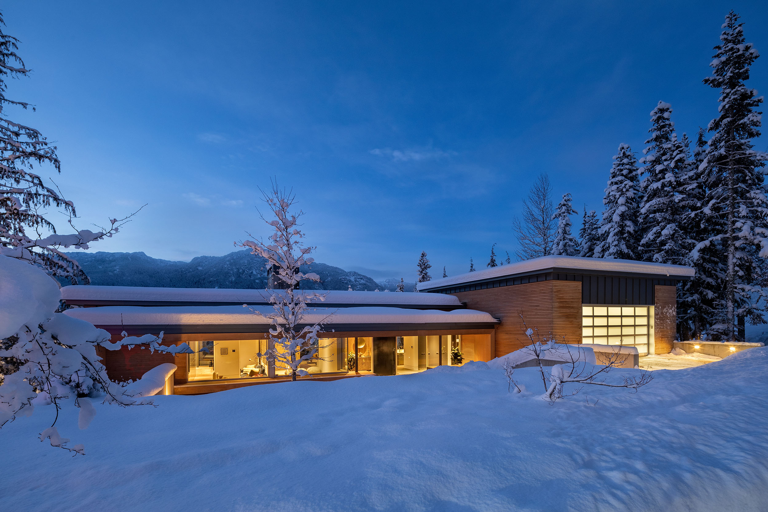 A Whistler home lit at dusk with snow-covered mountains behind